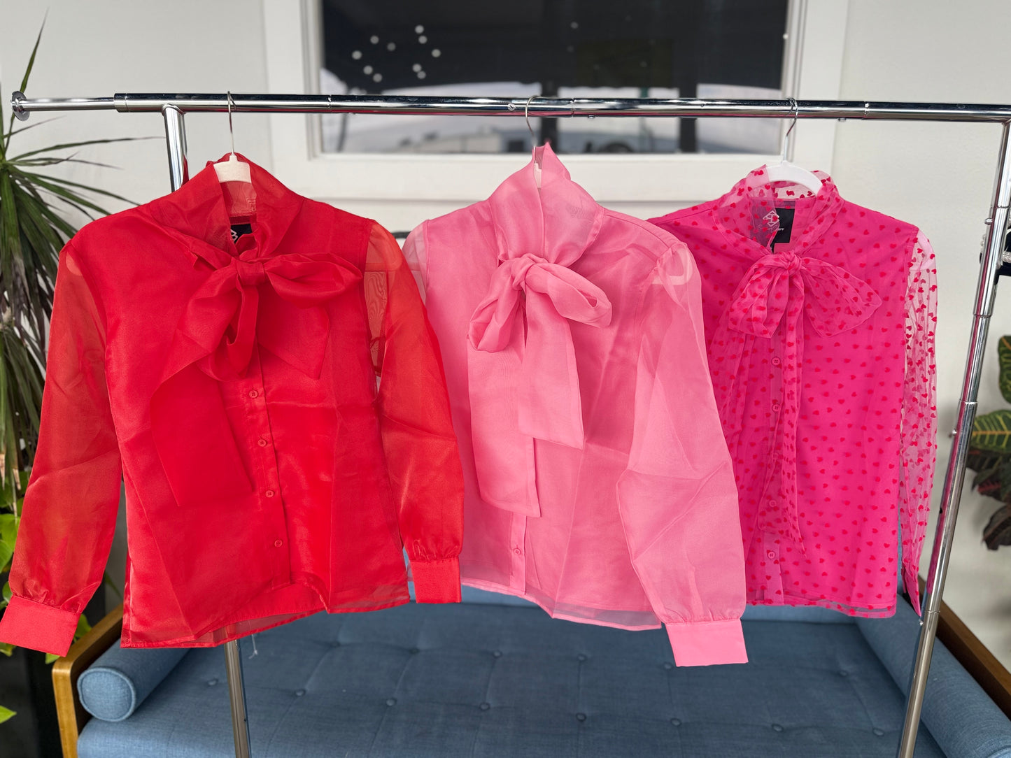 Three sheer blouses in red, pink, and polka dot, featuring bow ties and button closures, perfect for layering.