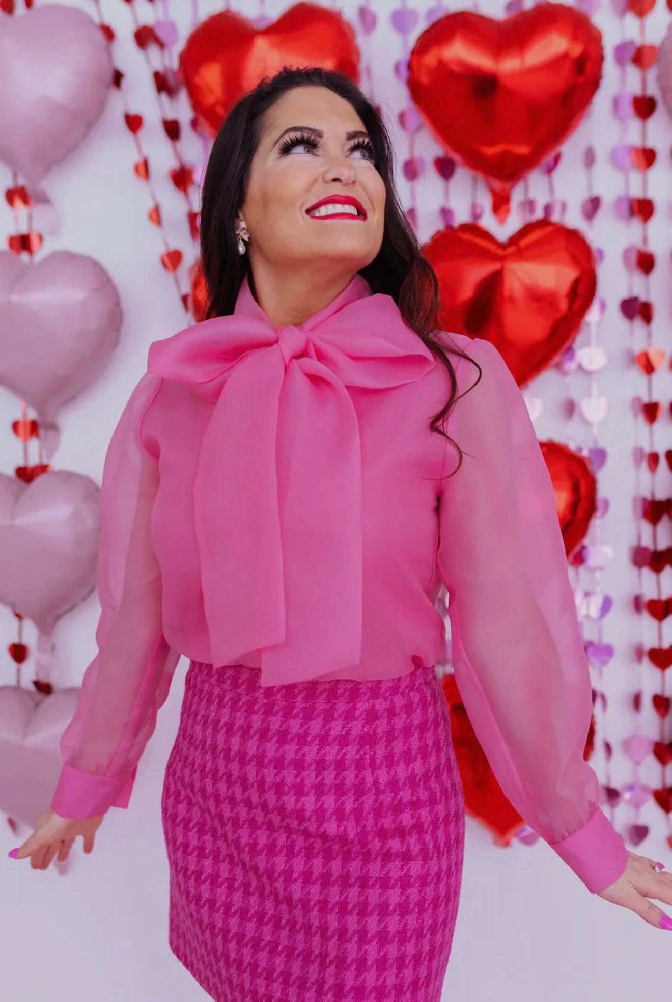 Pink sheer blouse with bow detail, paired with a pink houndstooth skirt, festive heart backdrop.
