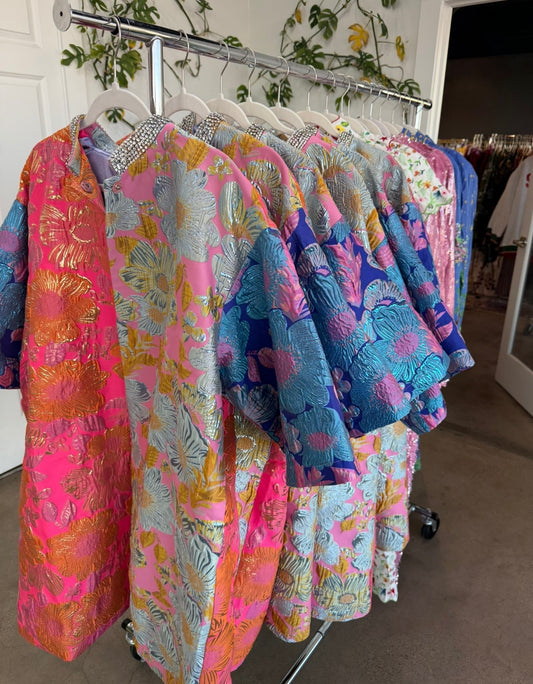 Colorful floral embroidered dresses in pink, blue, and gold on hangers, featuring stylish embellishments.